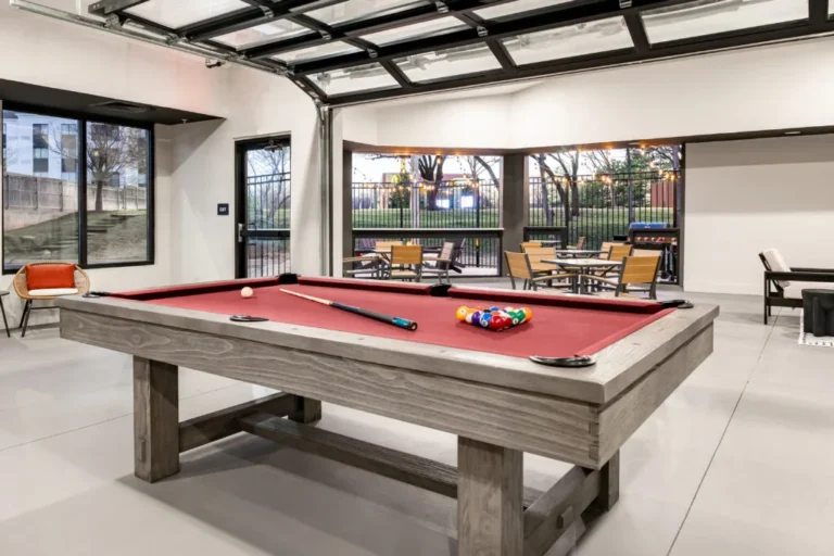 A pool table with a red felt surface, situated in a room with white walls and a large window offering a view of the outdoors.