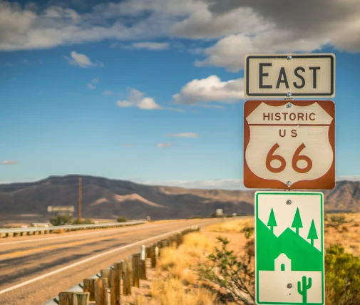 A vintage road sign reading "East" with a "66" in the middle, situated on the side of a desert highway.
