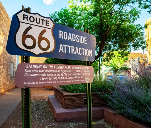 Sign with Route 66 logo and text about roadside attraction. Sign reads "STANDIN' ON THE CORNER PARK, WINSLOW, ARIZONA. This area was dedicated on September 11, 1999, to commemorate the memorable song of the 1970s that made Winslow, Arizona a town to sing about on famous Route 66. Recognized by Hampton Inn Save-A-Landmark program as one of 100 such sites.