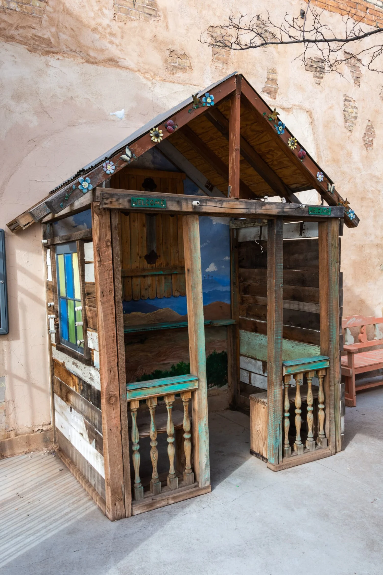 A small wooden structure with a sloping roof and open sides, featuring a bench and decorative elements, set against a stone wall.