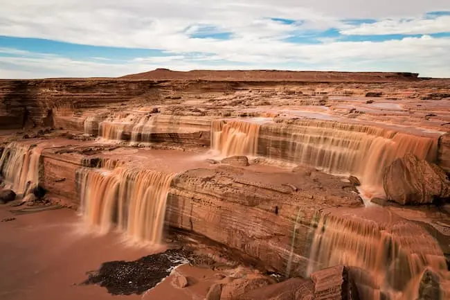 A large, red rock waterfall cascading into a river, with a vast, flat, red rock plateau in the background and a cloudy sky above.