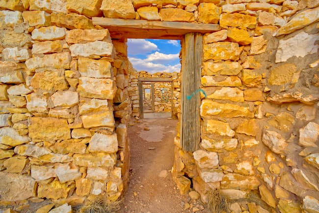 A stone wall with a doorway, featuring a wooden beam and a blue sky with white clouds in the background.