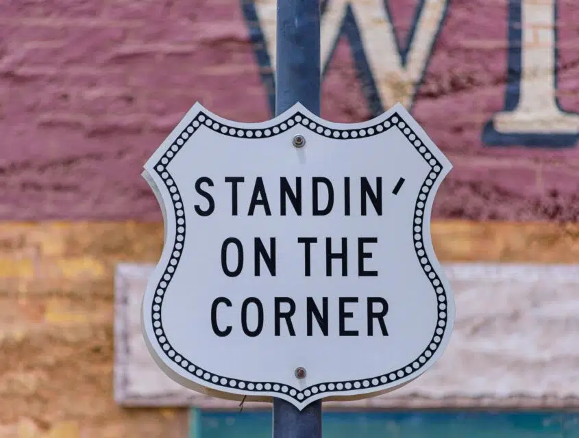 A sign with the text "STANDIN' ON THE CORNER" in black font, attached to a pole, with a blurred background featuring a red brick wall and a white sign with a blue border.