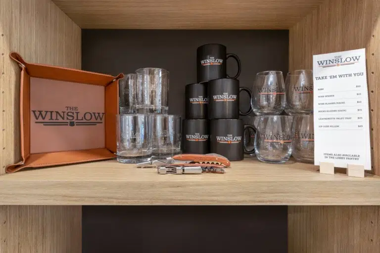 A wooden shelf displays a collection of Winslow, AZ branded merchandise, including a leather tray, coffee mugs, glasses, and a menu stand.