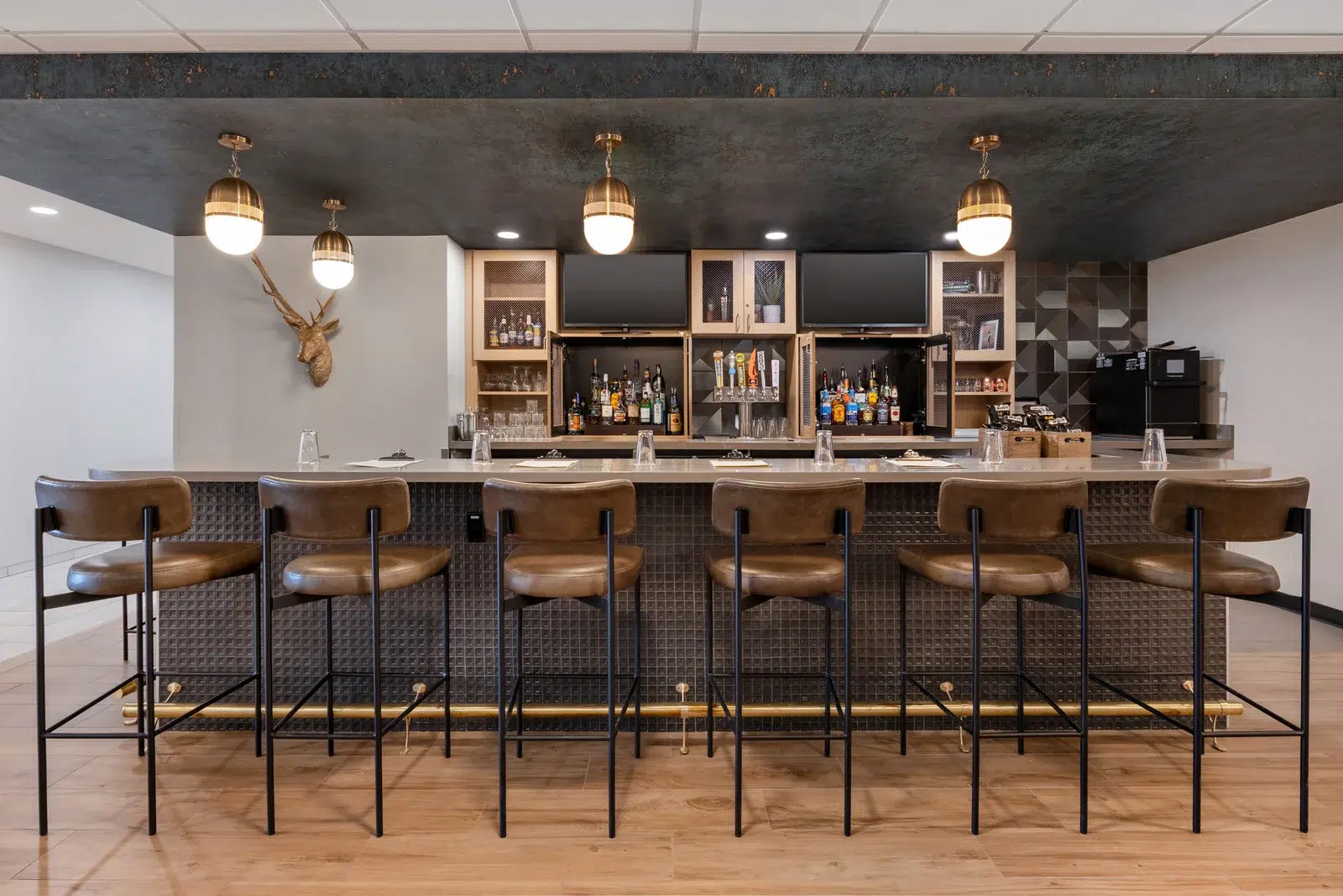 A modern bar with a white countertop, black metal frame, and brown leather stools, featuring a wall of shelves stocked with various bottles, a mounted deer head, and two lights hanging from the ceiling.