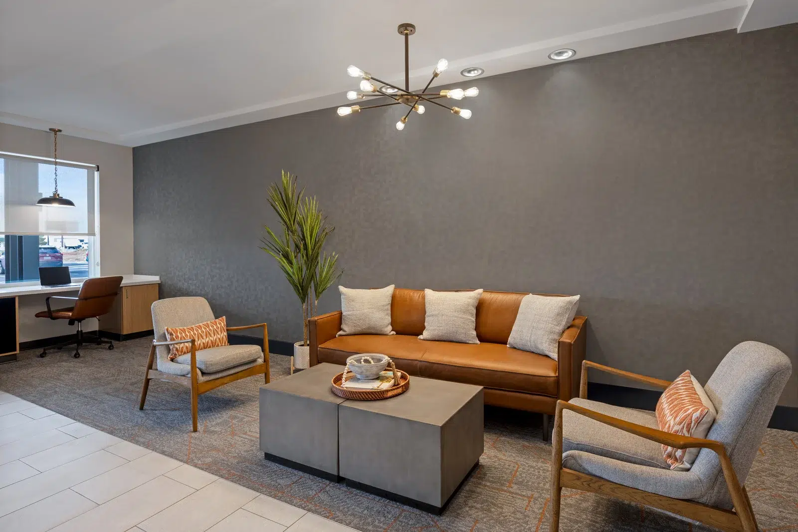 Modern living room with couch, chair, and coffee table. The couch is orange and the chair is gray. There is a plant in the corner and a desk in the background.