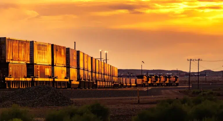 A train car with rusted metal panels and a flatbed, situated on a dirt track, with a vibrant orange sky in the background.