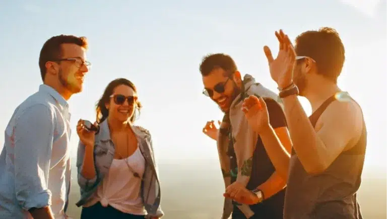 Three people standing outdoors, with one person waving at another, and the sun shining brightly behind them.