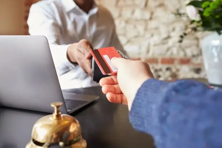 A person in a white shirt holds a credit card in front of a laptop computer and a golden bell.